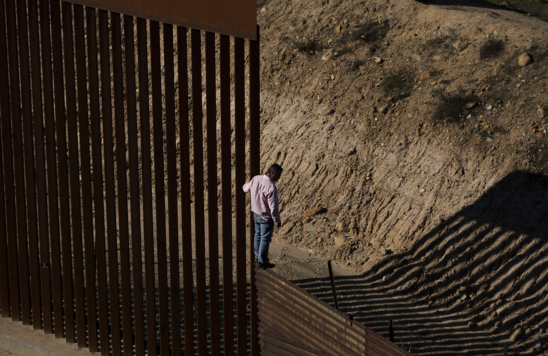 Un migrante mira hacia el suelo encaramado en lo alto de la valla fronteriza instantes antes de saltar para entrar a California desde Tijuana, México, el 28 de diciembre de 2018.