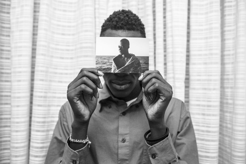 Malick poses for a photograph inside his room at the Hotel Colibri, holding a photo of himself taken on 2 August 2016, minutes after he was rescued from the waters of the Mediterranean Sea. He survived after seven hours of sailing from the coast of Libya, travelling in rubber dinghy with 120 people aboard.