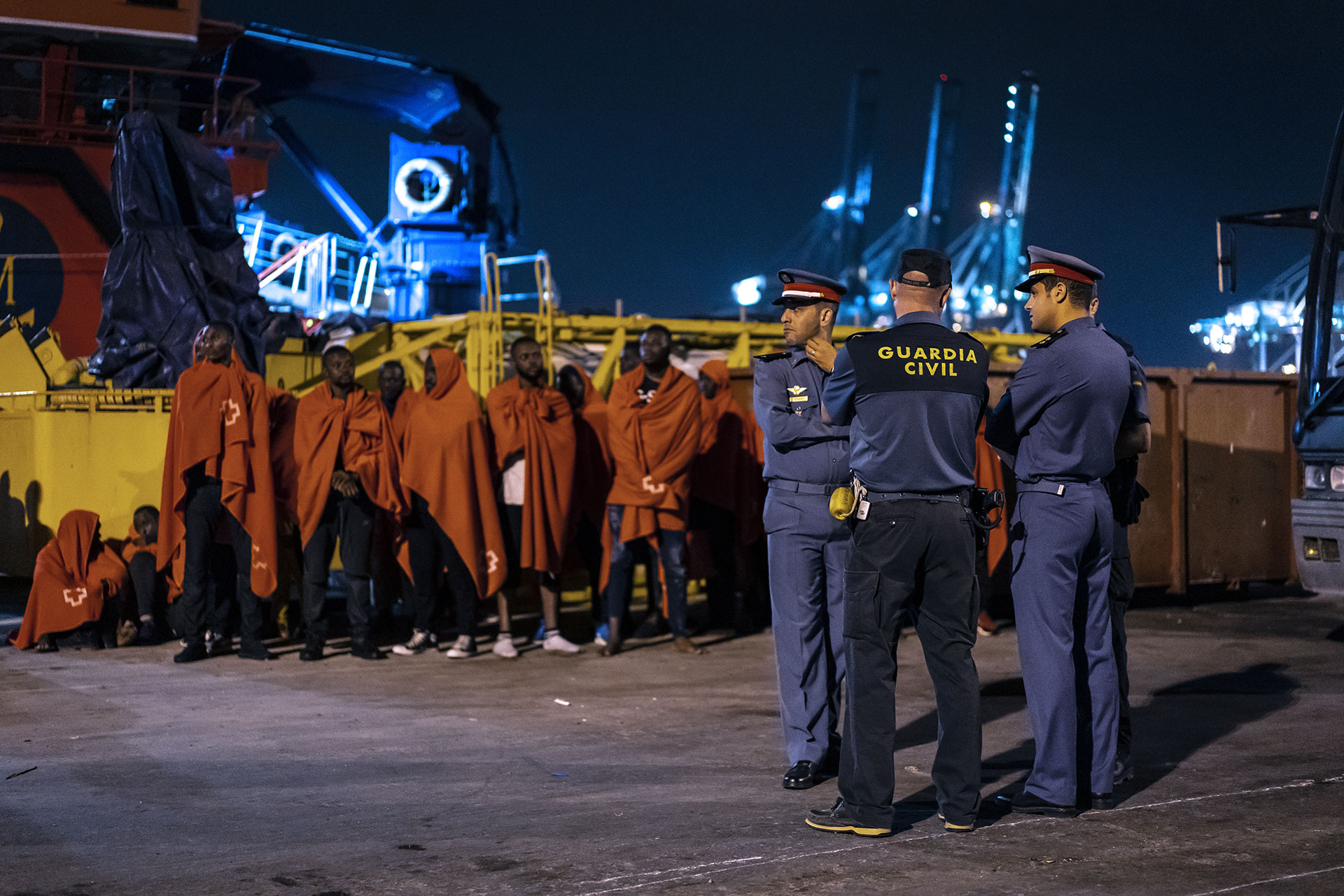 Algunas de las personas que fueron interceptadas por un barco de Salvamento Marítimo cuando intentaban llegar a la costa a bordo de un pequeño, esperan en el puerto de Algeciras cerca de una patrulla conjunta de Guardia Civil y Gendarmería Marroquí, 21 de Julio de 2019.