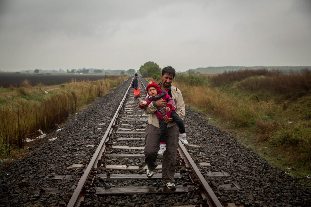 A father walks along the railroad tracks carrying his kid in his arms after having crossed the border between Serbia and Hungary. He is entering Roszke. (Roszke, Hungary. 10/09/2015).