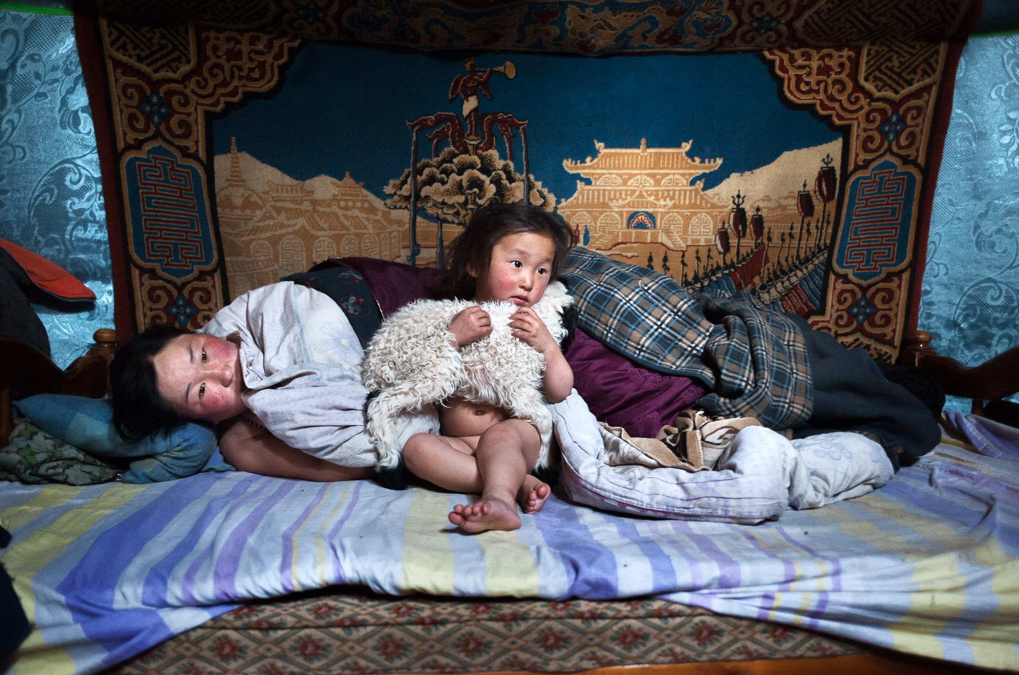 Mongolia, provincia de Arkhangay. Erdene Tuya, de 29 años, con su hijo de 3 años llamado Tuvchinj (abraza a un corderito que duerme con ellos). Acaban de despertarse mientras su marido Batgargal salió a echar un vistazo al rebaño con el otro hijo llamado Azjargal, de 6 años. En la provincia mongola de Arkhangai, la familia Tsamba vive al límite, luchando contra los duros inviernos junto a su rebaño de ovejas. Las severas condiciones invernales, conocidas como dzud, han sido las responsables de la muerte de la mitad del rebaño de la familia, que llegó a tener 2.000 ovejas, en los últimos tres inviernos. Recientemente, en busca de pastos más cálidos, los Tsambas se trasladaron desde la provincia de Bulgan, en el norte, a esta región cercana a un pueblo del centro de Mongolia llamado Ulziit.