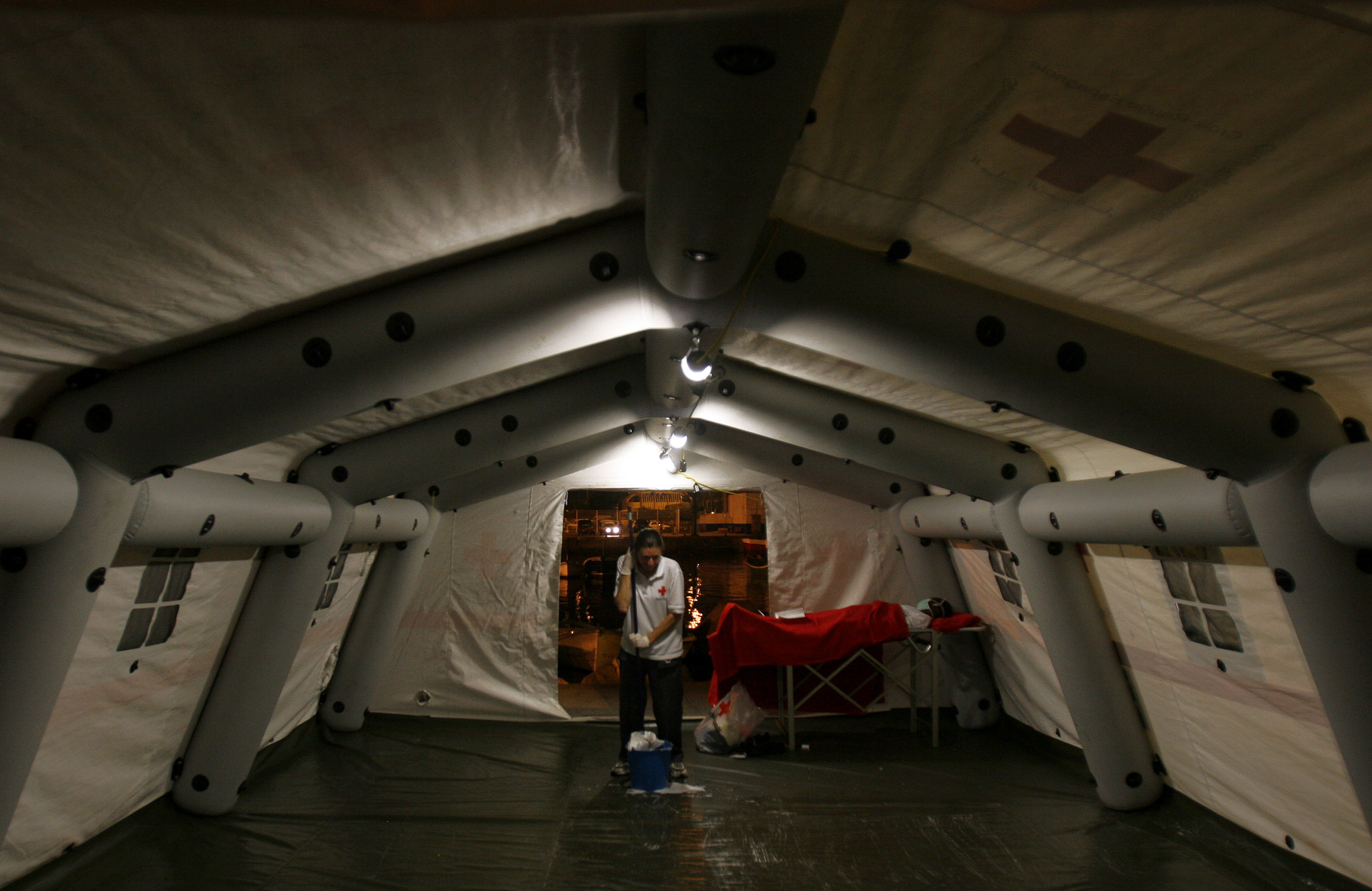 A Red Cross volunteer mops the floor of a tent where moments before a group of undocumented refugees arrived in dugout canoes at the port of Los Cristianos, Tenerife. One of them, weak from the trip, waits in a stretcher for the ambulance that will take him to the hospital. 17 August 2006.