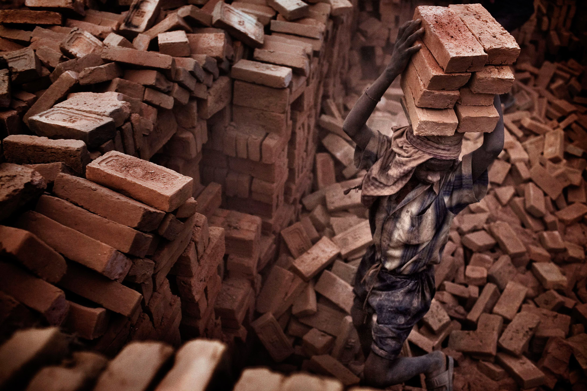 A young worker balances 8 bricks, each weighing 4 kilos, on his head. At every visit the brick pile he is exposed to significant quantities of hazardous dust.
