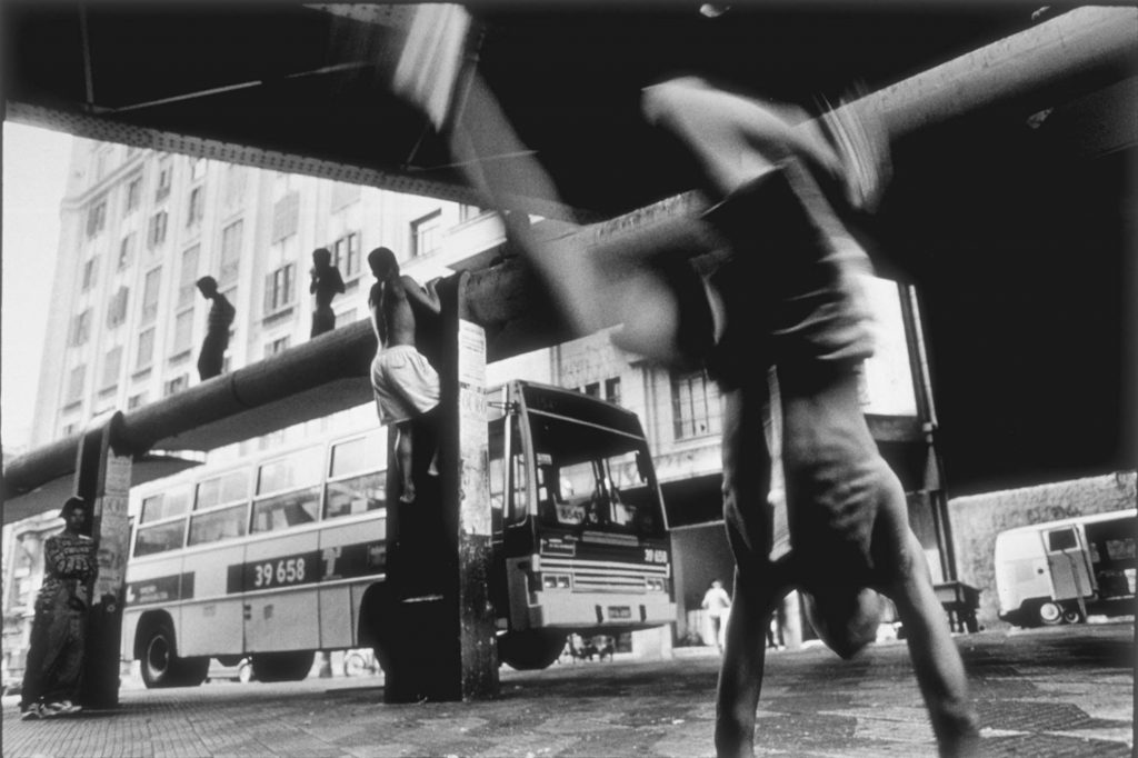 12 a. m. Niño en primer término practicamndo capoeira, arte marcial -danza típica de Brasil y originaria de África-. Al fondo un grupo de niños sobre una parada de autobús.