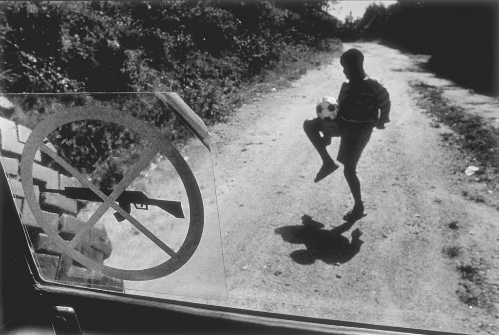 En la imagen, un niño juega al fútbol frente a la prohibición de portar armas que exhibe un blindado de la cooperación internacional. Junio, 1998.