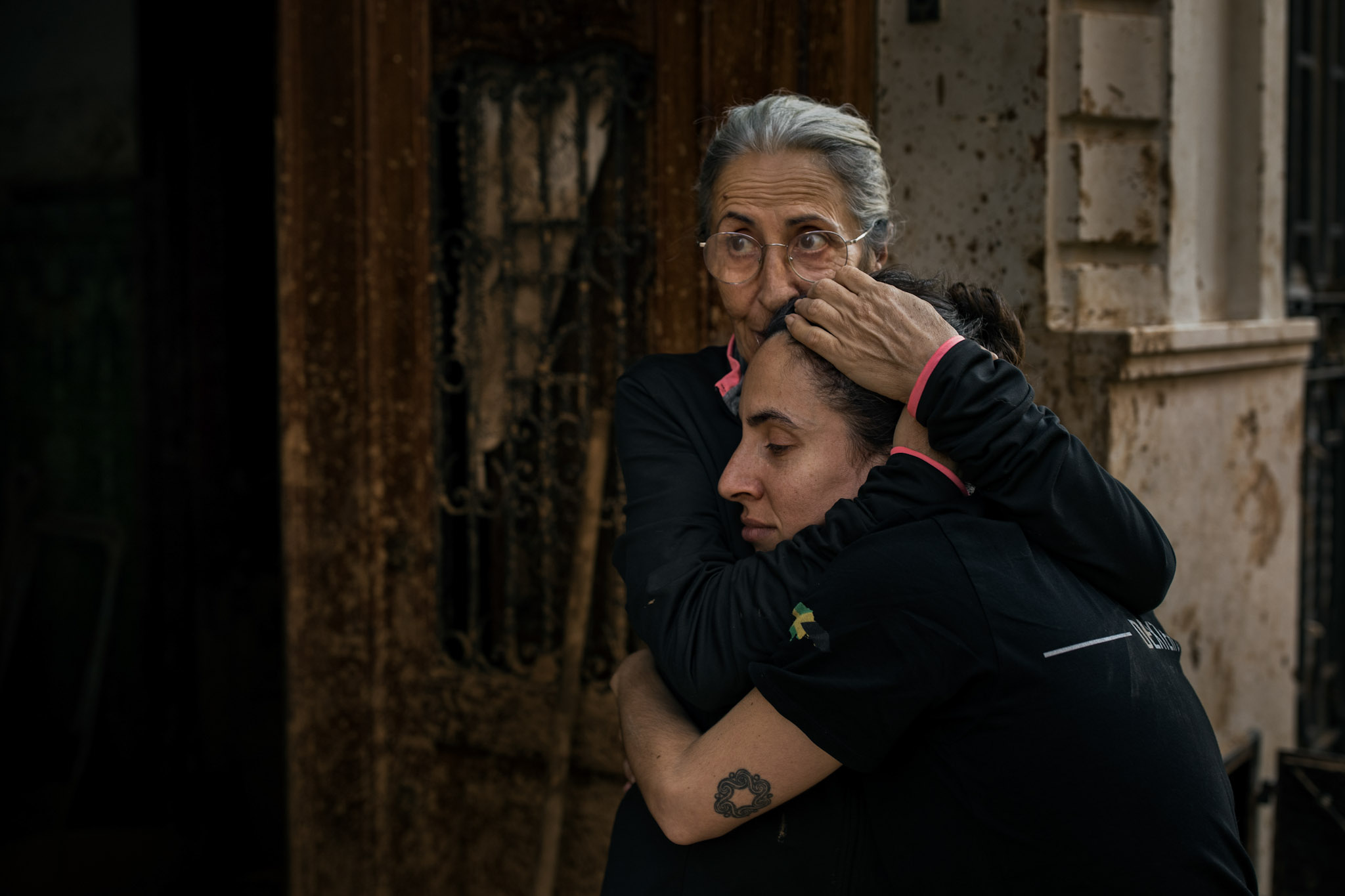 Agustina Zahonero del Río, de 61 años, y Gema, de 32, su hija mayor, se abrazan delante de la casa de Agustina, en Paiporta. Cuando la Generalitat Valenciana emitió la alerta por inundaciones, Agustina ya estaba agarrada a la lámpara del techo de su salón. El agua alcanzó casi tres metros en el interior de su vivienda, y tanto Agustina como Ana, de 26 años, su hija pequeña, flotaron en la oscuridad, durante horas, recibiendo los golpes de los objetos que arrastaba la riada. No fue hasta la madrugada cuando un vecino hizo un agujero en la pared con un martillo, lo que les permitió subir hasta la casa de la vecina del piso de arriba y ponerse a salvo.