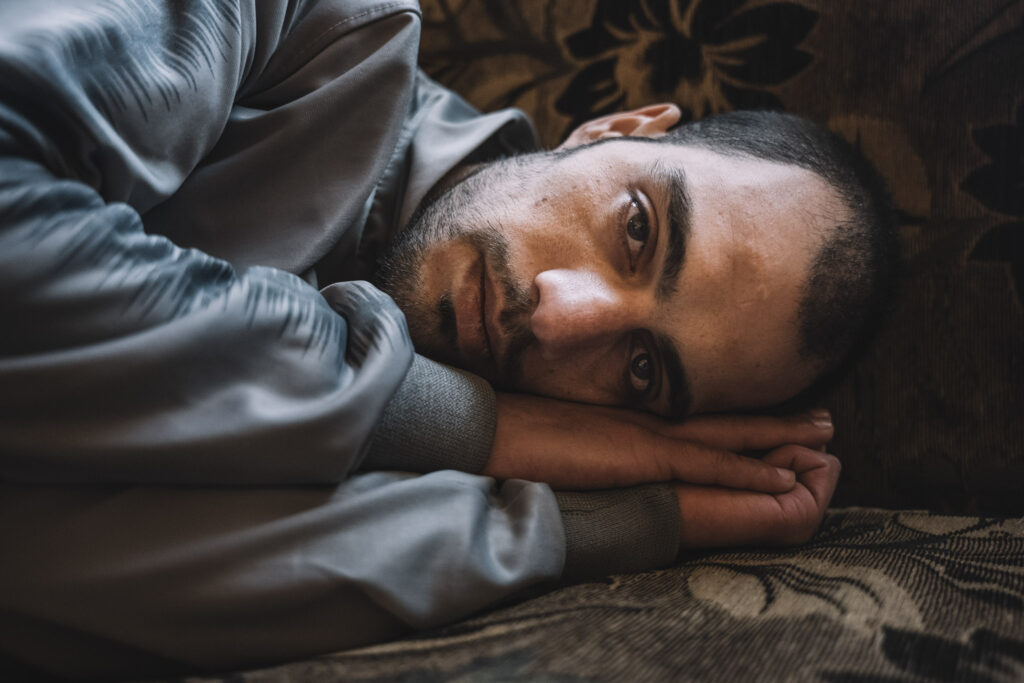 After seven years in prison, Mohamed Khaled Krayem is finally free. His family is happy, but he struggles to think about the future as he must attend medical visits to recover after his release. “They used to beat us with iron and plastic pipes on our hands and feet,” Mohamed says. Welcome: *ahlan wa sahlan.* The interviewees utter these words as if referring to an old acquaintance: *ahlan wa sahlan.* And another: *dulab* (wheel). This method of torture, though simple, became one of the regime's trademarks: seating the prisoner inside a wheel, restraining them, and beating them. “They put you on your back inside the tire and start hitting your feet with strips made from another tire they had cut,” explains Mohamed, the survivor with broken teeth. In the photo, Mohamed assumes the same position he used to sleep in while in prison. After seven years of lying in the same posture, his muscle memory, still fresh since his release only days ago, instinctively reproduces the positions he was forced to adopt in his cell. The lack of space and the repetitive nature of those movements mean he remembers exactly how he lay down and slept in Sednaya Jail.